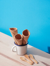 Three Empty Ice Cream Cones In A White Cup On A Blue Background With Wooden Spoons On A Wooden Table. Harsh Light With Shadows.
