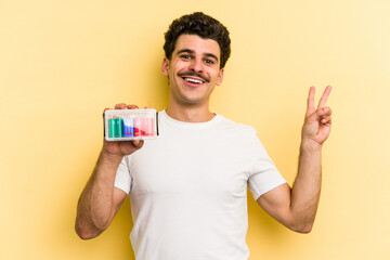 Young caucasian man holding batteries isolated on yellow background joyful and carefree showing a peace symbol with fingers.