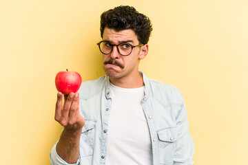 Young caucasian man holding an apple isolated on yellow background confused, feels doubtful and unsure.