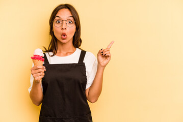 Wall Mural - Young hispanic dependent woman holding an ice-cream isolated on yellow background pointing to the side