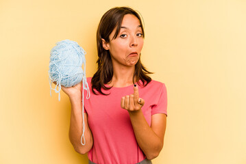 Wall Mural - Young hispanic woman holding a ball of wool isolated on yellow background pointing with finger at you as if inviting come closer.