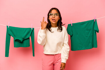 Wall Mural - Young hispanic woman holding laundry basket isolated on pink background showing a disappointment gesture with forefinger.