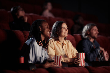 Two happy girls eating popcorn and laughing during watching comedy in the cinema