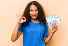 Young Hispanic Woman Holding Bank Notes Isolated On Beige Background Cheerful And Confident Showing Ok Gesture.