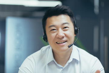 Close-up photo portrait of Asian man with video call headset, tech support worker smiling and looking into camera