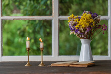 Two Burning Candles In Candlestick An Open Old Book And Bouquet Of Colorful Flowers On The Windowsill Next To The Window
