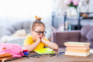 Cute thoughtful tired schoolgirl with glasses. Sitting at his desk at home and bored. Back to school. Little girl C does not want to get knowledge and do homework. Learning problems