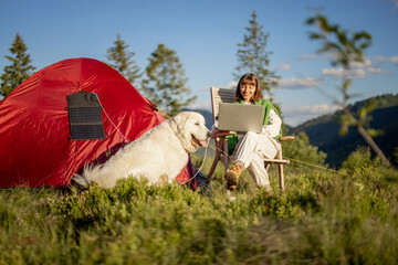 Young woman works on laptop while traveling with tent and dog in the mountains. Charging computer with portable solar panel hanging on tent. Concept of remote work at campsite