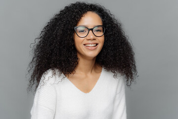 Wall Mural - Portrait of smiling African American woman wears transparent eyewear and white jumper, being in high spirit, expresses good emotions, isolated over grey background. People and ethnicity concept