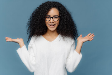 Wall Mural - Positive friendly young woman with Afro appearance, spreads palms, holds invisible object, has charming smile, healthy skin, wears spectacles and white sweater, isolated over blue background.