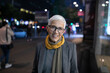 © Adam Radosavljevic - Smiling mature senior woman with short gray hair and eyeglasses walking on street, night scene in city