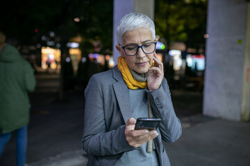 Wall Mural - Smiling mature senior woman with short gray hair and eyeglasses use phone on street, night scene in city