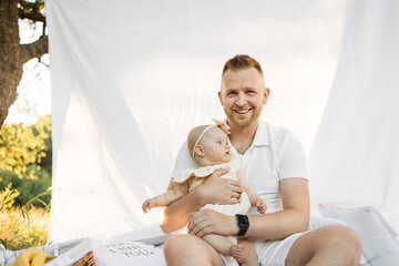 Wall Mural - Blond haired cheerful happy man relaxing on white blanket with curtain in park with little baby girl while looking at camera. Family summer concept.