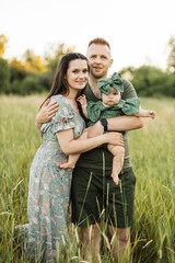 Wall Mural - Joyful young parents playing with cute daughter outdoors while standing on background of green meadow. Man and woman holding baby girl on hands. Copy space.