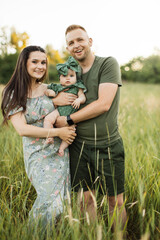 Wall Mural - Joyful young parents playing with cute daughter outdoors while standing on background of green meadow. Man and woman holding baby girl on hands. Copy space.