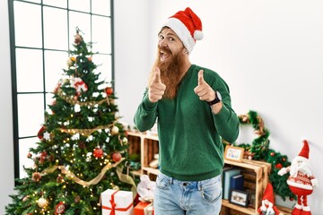 Sticker - Redhead man with long beard wearing christmas hat by christmas tree pointing fingers to camera with happy and funny face. good energy and vibes.