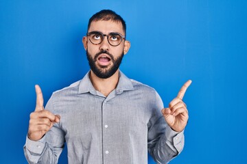 Wall Mural - Middle east man with beard standing over blue background amazed and surprised looking up and pointing with fingers and raised arms.