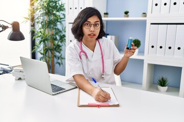 Canvas Print - Young latin woman wearing doctor uniform holding inhaler writing on clipboard at clinic