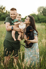 Wall Mural - Portrait of happy young parents embracing little cute daughter and their dog celebrate six months since the birth of a child while standing on the field on fresh air.