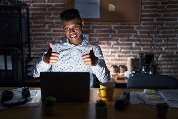 Wall Mural - Young hispanic man working at the office at night approving doing positive gesture with hand, thumbs up smiling and happy for success. winner gesture.