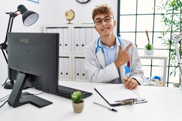 Canvas Print - Young caucasian doctor man working at the clinic cheerful with a smile of face pointing with hand and finger up to the side with happy and natural expression on face