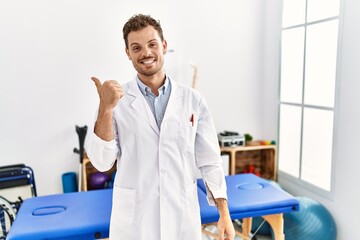 Wall Mural - Handsome young man working at pain recovery clinic smiling with happy face looking and pointing to the side with thumb up.