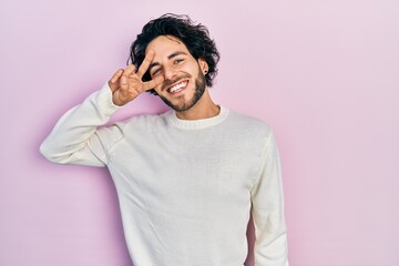 Poster - Handsome hispanic man wearing casual white sweater doing peace symbol with fingers over face, smiling cheerful showing victory