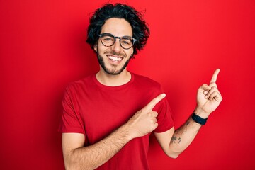 Wall Mural - Handsome hispanic man wearing casual t shirt and glasses smiling and looking at the camera pointing with two hands and fingers to the side.