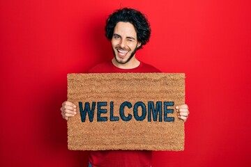 Canvas Print - Handsome hispanic man holding welcome doormat winking looking at the camera with sexy expression, cheerful and happy face.