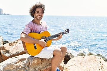 Canvas Print - Young hispanic man playing classical guitar sitting on rock at the beach.