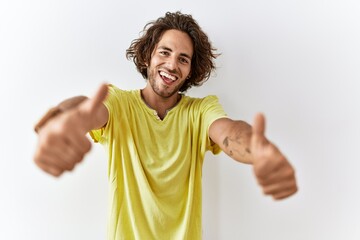 Wall Mural - Young hispanic man standing over isolated background approving doing positive gesture with hand, thumbs up smiling and happy for success. winner gesture.