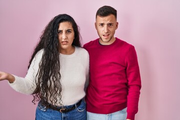 Sticker - Young hispanic couple standing over pink background in shock face, looking skeptical and sarcastic, surprised with open mouth