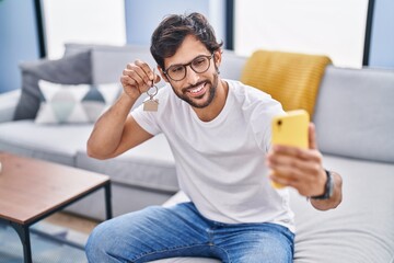 Wall Mural - Young hispanic man make selfie by smartphone holding key at home