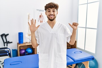 Sticker - Young arab man working at pain recovery clinic showing and pointing up with fingers number six while smiling confident and happy.