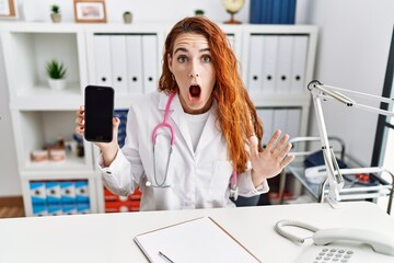 Sticker - Young redhead doctor woman holding smartphone showing blank screen scared and amazed with open mouth for surprise, disbelief face