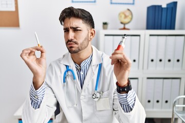 Poster - Young doctor man holding electronic cigarette at medical clinic clueless and confused expression. doubt concept.