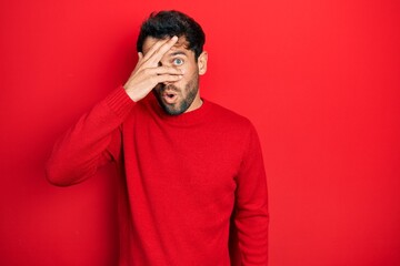 Canvas Print - Handsome man with beard wearing casual red sweater peeking in shock covering face and eyes with hand, looking through fingers with embarrassed expression.