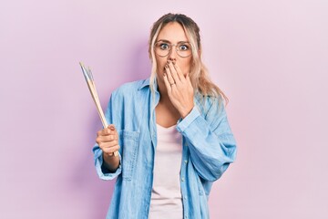 Poster - Beautiful young blonde woman holding paintbrushes covering mouth with hand, shocked and afraid for mistake. surprised expression