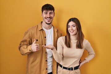 Young hispanic couple standing over yellow background showing palm hand and doing ok gesture with thumbs up, smiling happy and cheerful