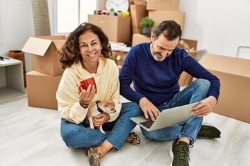 Poster - Middle age hispanic couple using laptop and drinking coffee. Sitting on the floor with dog at new home.