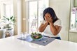 Young hispanic woman eating healthy salad at home with sad expression covering face with hands while crying. depression concept.