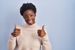 © Krakenimages.com - African american woman standing over blue background success sign doing positive gesture with hand, thumbs up smiling and happy. cheerful expression and winner gesture.