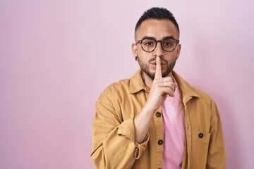 Sticker - Young hispanic man standing over pink background asking to be quiet with finger on lips. silence and secret concept.