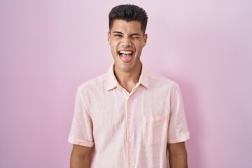 Wall Mural - Young hispanic man standing over pink background sticking tongue out happy with funny expression. emotion concept.