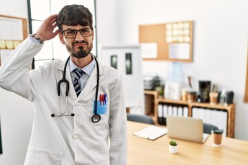 Sticker - Hispanic man with beard wearing doctor uniform and stethoscope at the office confuse and wonder about question. uncertain with doubt, thinking with hand on head. pensive concept.