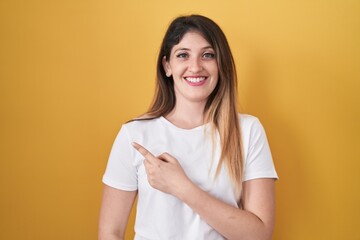 Poster - Young brunette woman standing over yellow background cheerful with a smile of face pointing with hand and finger up to the side with happy and natural expression on face