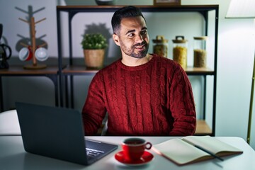 Wall Mural - Young hispanic man with beard using computer laptop at night at home looking away to side with smile on face, natural expression. laughing confident.