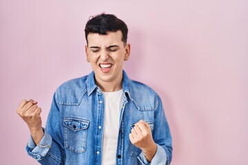 Wall Mural - Non binary person standing over pink background very happy and excited doing winner gesture with arms raised, smiling and screaming for success. celebration concept.