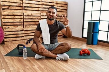 Poster - Young indian man sitting on training mat at the gym smiling looking to the camera showing fingers doing victory sign. number two.