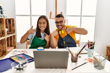 Poster - Young hispanic couple using laptop sitting on the table at art studio looking confident with smile on face, pointing oneself with fingers proud and happy.